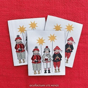 Könnte beinhalten: Drei weiße Stoffetiketten mit gestickten Illustrationen von Kindern in Winterkleidung, jeweils unter einem goldenen Stern. Die Kinder tragen rote, graue und schwarze Mäntel, Mützen und Hosen. Der Text "acufactum UTE MENZE" befindet sich unten.