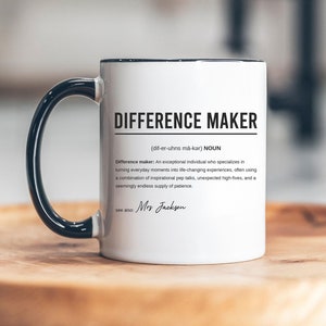 Puede incluir: Taza de café de cerámica blanca con asa negra. La taza tiene un borde negro alrededor de la parte superior y el texto "DIFFERENCE MAKER (dif-er-uhns mä-kar) NOUN" impreso en negro. El texto continúa con una definición de la palabra "difference maker" y termina con el texto "ver también: Mrs Jackson".