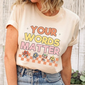 May include: A cream colored t-shirt with the text "YOUR WORDS MATTER" in pink, yellow, and orange. The text is surrounded by a checkered pattern and cartoon characters.
