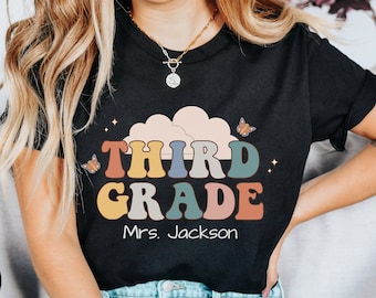 May include: Black t-shirt with a retro-style graphic that says "Third Grade" in colorful letters. The graphic also includes a cloud and butterflies. The text "Mrs. Jackson" is below the graphic.