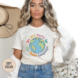 May include: A woman wearing a cream colored t-shirt with a graphic of a blue globe with smiling faces and the text "We All Smile In The Same Language".