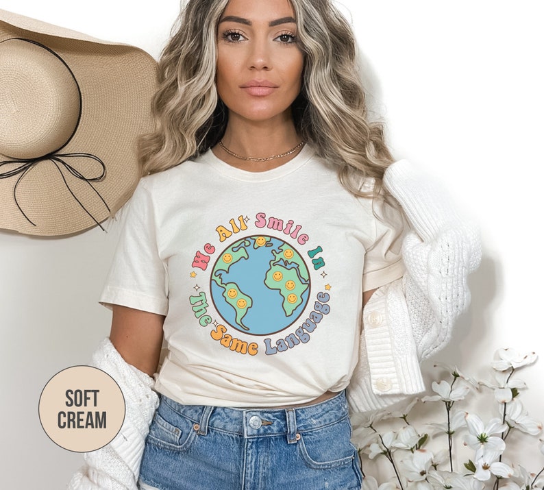 May include: A woman wearing a cream colored t-shirt with a graphic of a blue globe with smiling faces and the text "We All Smile In The Same Language".