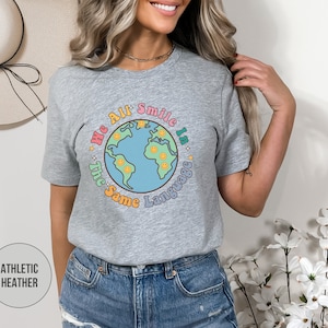May include: A heather grey t-shirt with a graphic of a colorful earth with smiley faces and the text "We All Smile In The Same Language".