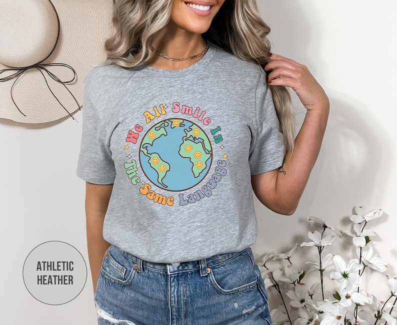 May include: A heather grey t-shirt with a graphic of a colorful earth with smiley faces and the text "We All Smile In The Same Language".
