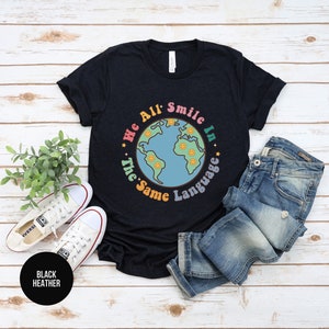 May include: A black heather t-shirt with a graphic of a blue and green earth with smiling faces around it. The text "We All Smile In The Same Language" is written around the earth.