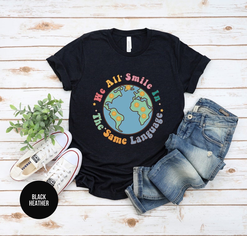 May include: A black heather t-shirt with a graphic of a blue and green earth with smiling faces around it. The text "We All Smile In The Same Language" is written around the earth.