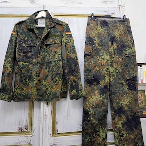 Puede incluir: Conjunto de chaqueta y pantalón de camuflaje militar. La chaqueta, con cuatro bolsillos y cuello, y el pantalón, con múltiples bolsillos, presentan un patrón verde, marrón y negro. La bandera alemana es visible en la manga de la chaqueta.