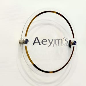 May include: A clear acrylic sign with a gold ring around the edge. The sign says "Aeym's Studio" in black text.