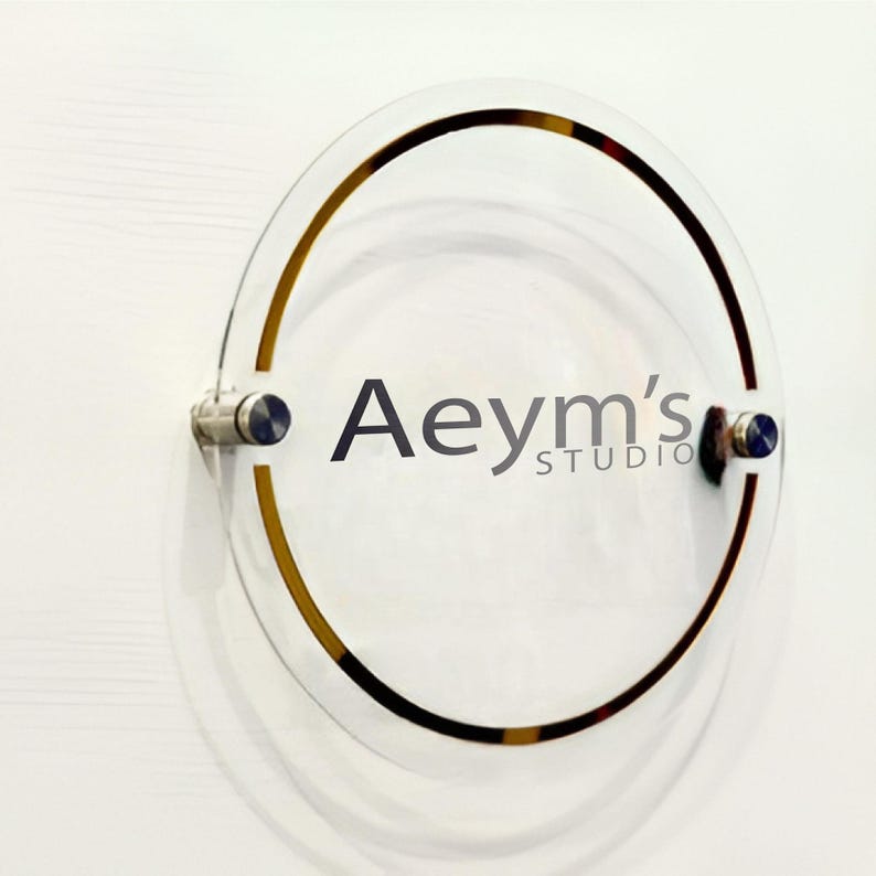 May include: A clear acrylic sign with a gold ring around the edge. The sign says "Aeym's Studio" in black text.