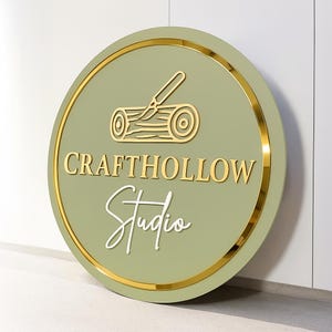 Może przedstawiać: Okrągły znak z szałwiowym zielonym tłem i złotą obw&oacute;dką. Znak zawiera słowa "CRAFTHOLLOW Studio" w złotych i białych literach, a także złotą ilustrację kłody i noża do rzeźbienia.