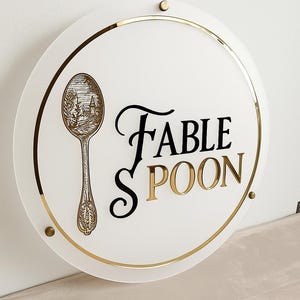 May include: A round white sign with gold trim and accents. The sign features the words "FABLE SPOON" in black and gold lettering. A detailed illustration of a spoon with a landscape scene is on the left side of the sign.
