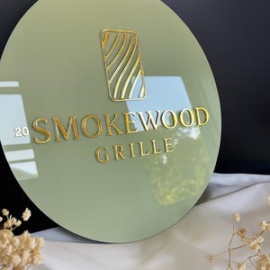May include: A round, sage green sign with gold lettering and a gold graphic. The sign reads "20 SMOKEWOOD GRILLE". The graphic is a gold rectangle with gold wavy lines inside. The sign is displayed on a white cloth.