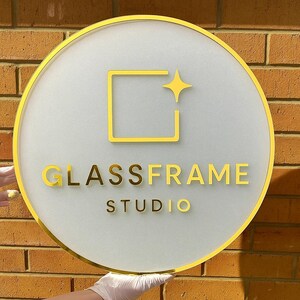 May include: A round sign with a gold border and the words "GLASSFRAME STUDIO" in gold lettering. The sign has a frosted glass appearance with a gold square and star design. The sign is held in front of a brick wall.