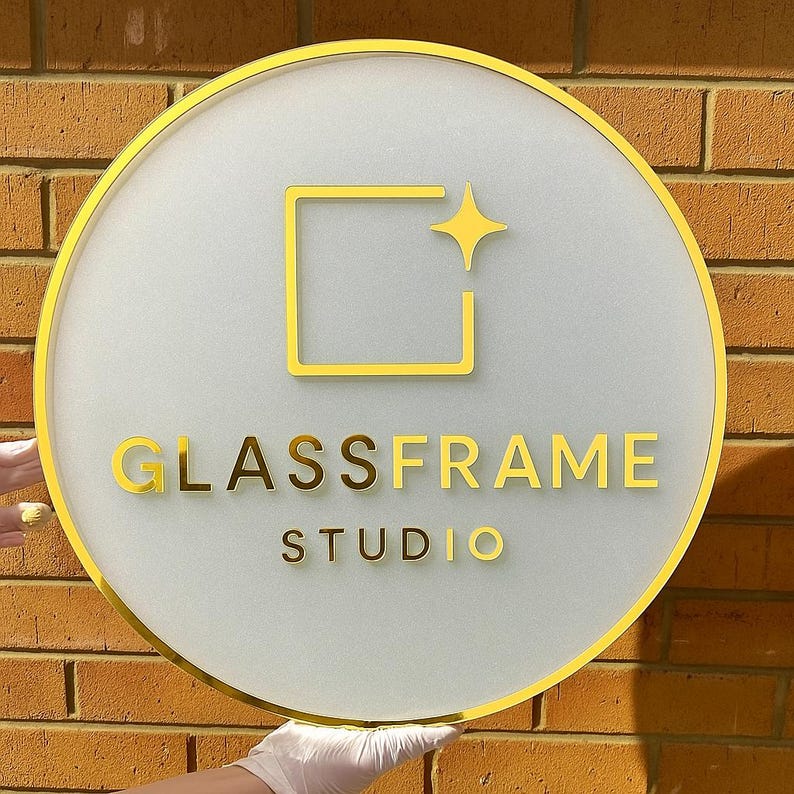 May include: A round sign with a gold border and the words "GLASSFRAME STUDIO" in gold lettering. The sign has a frosted glass appearance with a gold square and star design. The sign is held in front of a brick wall.