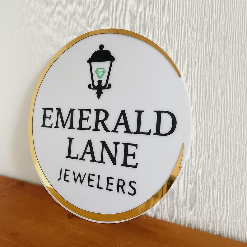 May include: A round white sign with a gold border and the words "EMERALD LANE JEWELERS" in black. The sign features a black lamp post icon with a green diamond shape inside.