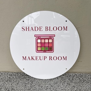 May include: A white, round sign with the words "SHADE BLOOM" and "MAKEUP ROOM" in burgundy. A pink and burgundy makeup palette graphic is centered between the text. The sign has four holes for mounting.