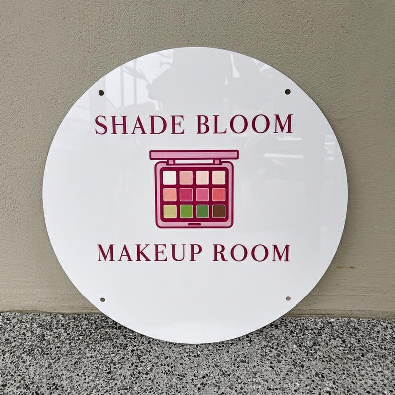 May include: A white, round sign with the words "SHADE BLOOM" and "MAKEUP ROOM" in burgundy. A pink and burgundy makeup palette graphic is centered between the text. The sign has four holes for mounting.