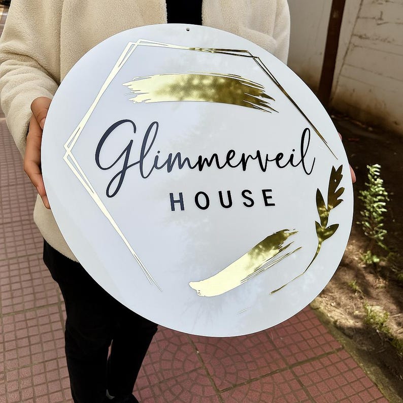 Pode incluir: Uma placa redonda branca com o texto "Glimmerveil HOUSE" em letras pretas cursivas e de bloco. A placa apresenta detalhes dourados, incluindo uma moldura geom&eacute;trica e designs de pinceladas. Um design de folha dourada est&aacute; &agrave; direita.