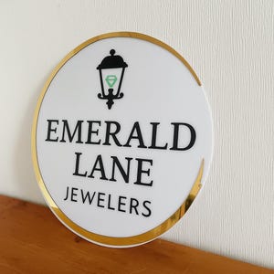 Puede incluir: Un letrero circular blanco con un borde dorado que presenta un gr&aacute;fico negro de una linterna que contiene una forma de diamante verde. El texto EMERALD LANE JEWELERS est&aacute; impreso en letras may&uacute;sculas negras, negritas y sin remates.