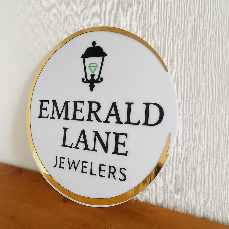 Puede incluir: Un letrero circular blanco con un borde dorado que presenta un gr&aacute;fico negro de una linterna que contiene una forma de diamante verde. El texto EMERALD LANE JEWELERS est&aacute; impreso en letras may&uacute;sculas negras, negritas y sin remates.