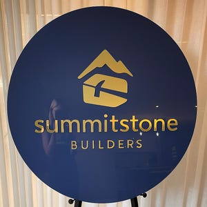 Puede incluir: Un letrero circular azul oscuro con un logotipo dorado de un pico de monta&ntilde;a sobre un icono de martillo. El texto "summitstone" aparece en letras min&uacute;sculas doradas, con la palabra "BUILDERS" en may&uacute;sculas doradas debajo.