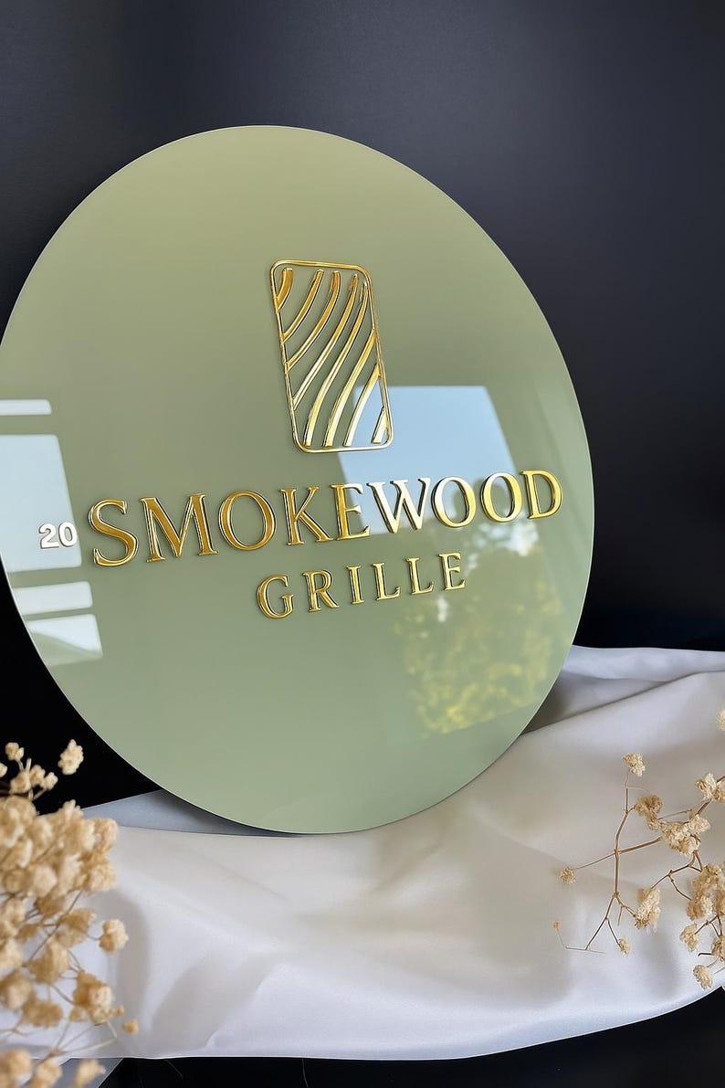 May include: A round, sage green sign with gold lettering and a gold graphic. The sign reads "20 SMOKEWOOD GRILLE". The graphic is a rectangular shape with gold wavy lines inside. The sign is displayed on a white cloth.