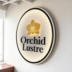 Może przedstawiać: Owalny znak z napisem "Orchid Lustre" w kolorze czarnym, pod złotym brokatowym wzorem orchidei. Znak ma białe tło z czarną obw&oacute;dką i jest zamontowany na białej ścianie.