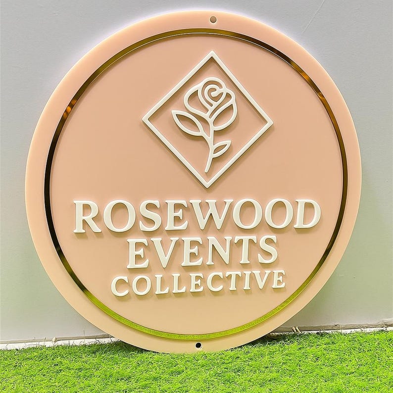 May include: A round sign with a peach-colored background and gold trim. The sign features the text "ROSEWOOD EVENTS COLLECTIVE" in white, capital letters. A white rose logo is centered above the text. The sign has two small holes at the top for hanging.