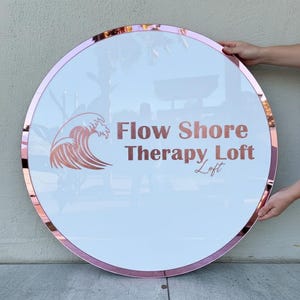 May include: A round sign with a white center and a rose gold border. The sign features the words "Flow Shore Therapy Loft" in rose gold lettering, along with a wave graphic. The sign is held by two hands.