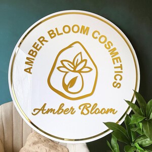 May include: A round white sign with gold trim and lettering. The sign reads "AMBER BLOOM COSMETICS" around the top and "Amber Bloom" in cursive at the bottom. A gold logo of a stylized flower is in the center.