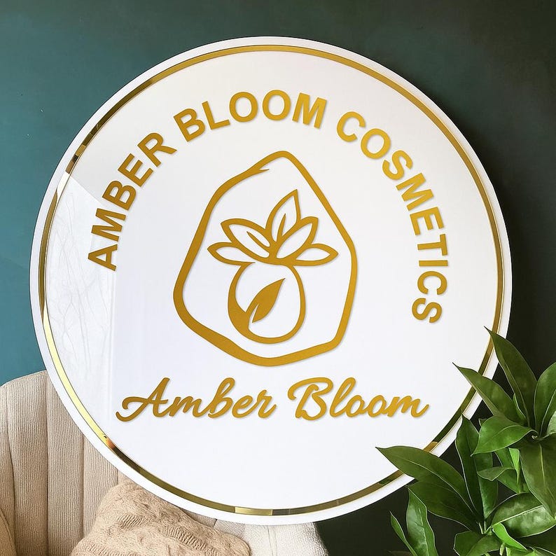 May include: A round white sign with gold trim and lettering. The sign reads "AMBER BLOOM COSMETICS" around the top and "Amber Bloom" in cursive at the bottom. A gold logo of a stylized flower is in the center.