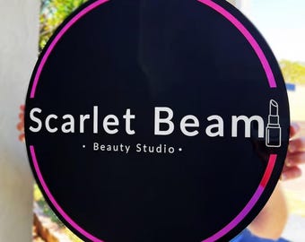 Può includere: Un cartello rotondo nero con un bordo rosa e magenta. Il cartello recita "Scarlet Beam" in testo bianco, con "Beauty Studio" sotto. Un'icona di rossetto è a destra.
