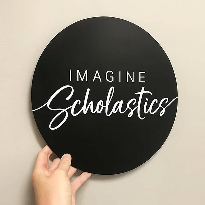 May include: A black, round sign with the words "IMAGINE Scholastics" in white lettering. The sign is held by a hand against a neutral background. The word "Imagine" is in a sans-serif font, and "Scholastics" is in a cursive font.