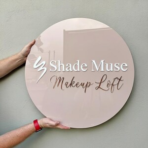 May include: A round, blush-colored sign with the text "Shade Muse Makeup Loft" in white and gold lettering. The sign is held by two hands, and a red watch is visible on one wrist. The sign has a glossy finish.