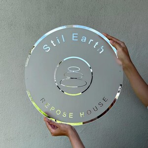 May include: A round, frosted glass sign with the words "Stil Earth" and "Repose House" in a light gray font. The sign features a silver-colored emblem of stacked stones. The sign is held by two hands.