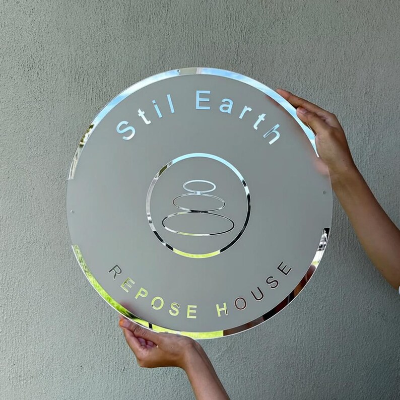 May include: A round, frosted glass sign with the words "Stil Earth" and "Repose House" in a light gray font. The sign features a silver-colored emblem of stacked stones. The sign is held by two hands.