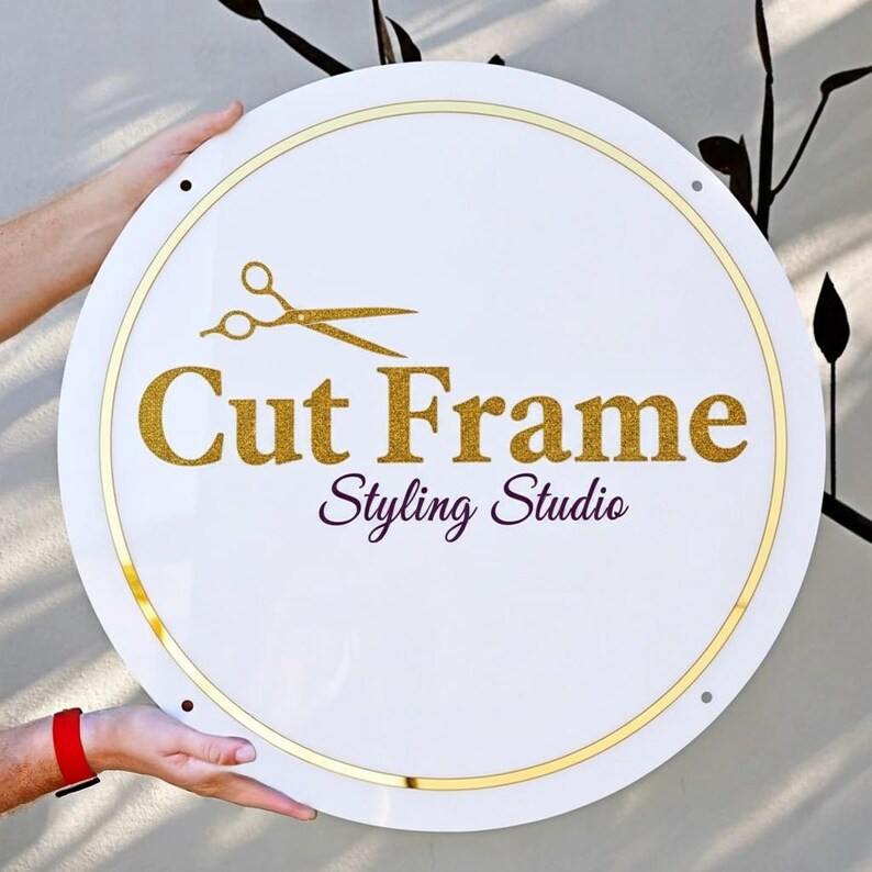 May include: A round white sign with gold trim and the words "Cut Frame Styling Studio" in gold glitter lettering. A gold scissors graphic is above the text. The sign is held by two hands.