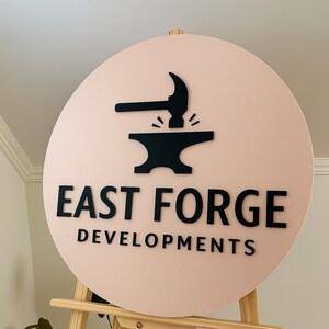 Puede incluir: Un letrero circular con fondo rosa p&aacute;lido que presenta un logotipo negro en 3D de un martillo golpeando un yunque. Debajo del logo, el texto negro dice EAST FORGE DEVELOPMENTS en una fuente sans-serif. El letrero est&aacute; sobre un caballete.