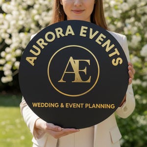 Pode incluir: Uma placa circular preta com letras douradas. Exibe "AURORA EVENTS" no topo e "WEDDING & EVENT PLANNING" na parte inferior. O centro apresenta um logotipo dourado com as letras "AE" dentro de um c&iacute;rculo dourado.