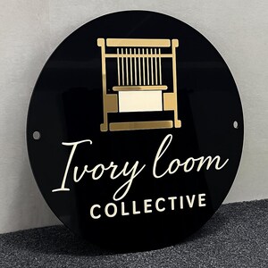 Puede incluir: Un letrero circular negro que presenta un gr&aacute;fico de color dorado de un telar en la parte superior. Debajo del gr&aacute;fico, el texto "Ivory loom" est&aacute; escrito en cursiva dorada, seguido de la palabra "COLLECTIVE" en letras may&uacute;sculas doradas.