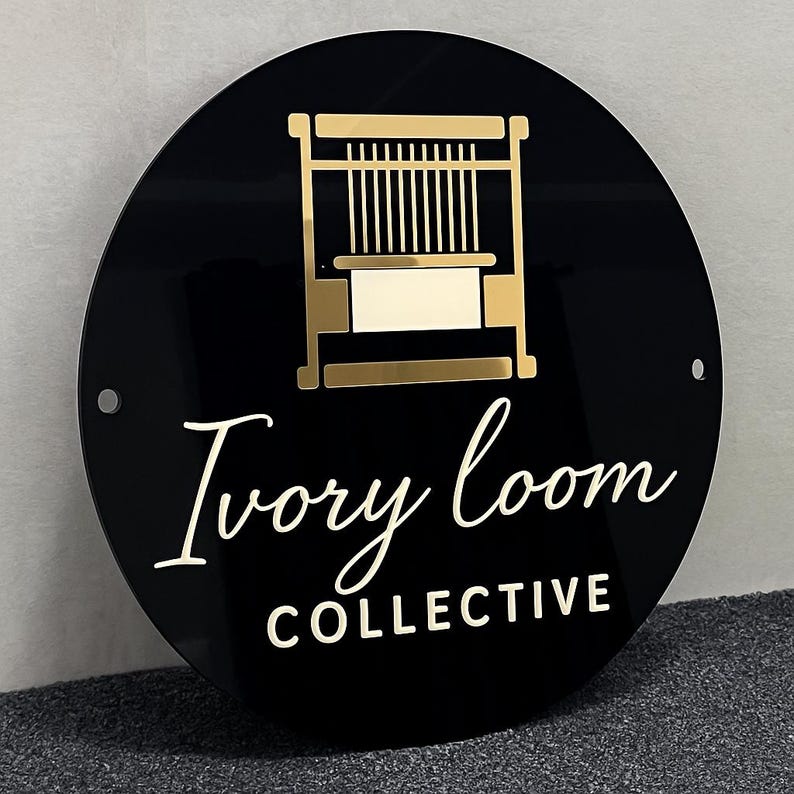 Puede incluir: Un letrero circular negro que presenta un gr&aacute;fico de color dorado de un telar en la parte superior. Debajo del gr&aacute;fico, el texto "Ivory loom" est&aacute; escrito en cursiva dorada, seguido de la palabra "COLLECTIVE" en letras may&uacute;sculas doradas.