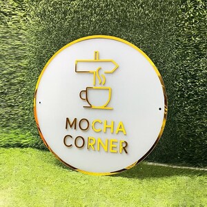 Może przedstawiać: Okrągły, biały znak ze złotą obw&oacute;dką i złotą grafiką o tematyce kawowej. Znak zawiera napis "MOCHA CORNER" złotymi literami. Tło jest zielone.