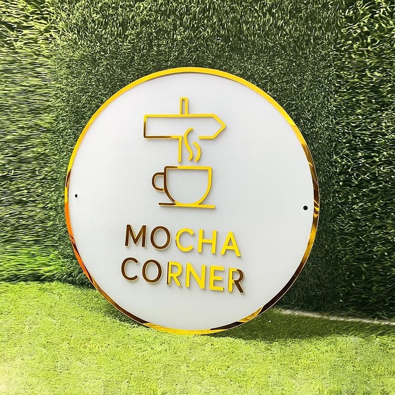 Może przedstawiać: Okrągły, biały znak ze złotą obw&oacute;dką i złotą grafiką o tematyce kawowej. Znak zawiera napis "MOCHA CORNER" złotymi literami. Tło jest zielone.