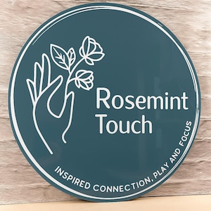 May include: A round sign with a teal background and white text and graphics. The text reads "Rosemint Touch" with the words "Inspired Connection, Play and Focus" around the bottom edge. A white hand holds a flower.