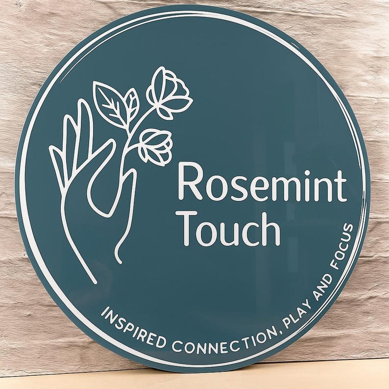 May include: A round sign with a teal background and white text and graphics. The text reads "Rosemint Touch" with the words "Inspired Connection, Play and Focus" around the bottom edge. A white hand holds a flower.