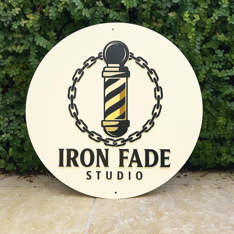 May include: A round, cream-colored sign with the words "IRON FADE STUDIO" in black lettering. The sign features a barber pole design in black and gold, encircled by a black chain. The sign is displayed against a backdrop of green foliage.