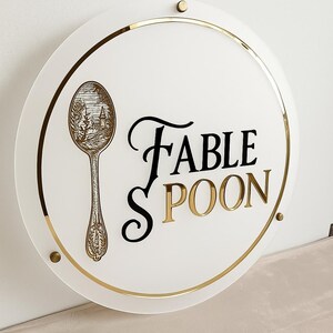 May include: A round white sign with gold trim and accents. The sign features the words "FABLE SPOON" in black and gold lettering. A detailed illustration of a spoon with a landscape scene is on the left side of the sign.