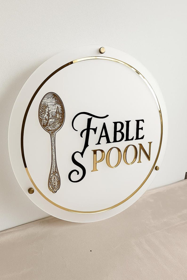 May include: A round white sign with gold trim and accents. The sign features the words "FABLE SPOON" in black and gold lettering. A detailed illustration of a spoon with a landscape scene is on the left side of the sign.