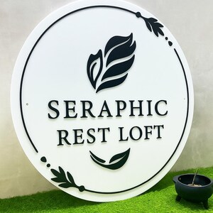 Pode incluir: Uma placa redonda branca com texto preto e motivos de folhas. A placa diz "SERAPHIC REST LOFT" em uma fonte sem serifa. Desenhos de folhas e pontos pretos circundam a placa. Um pequeno vaso preto com uma planta est&aacute; por perto.