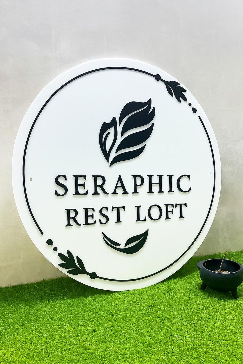 Pode incluir: Uma placa redonda branca com texto preto e motivos de folhas. A placa diz "SERAPHIC REST LOFT" em uma fonte sem serifa. Desenhos de folhas e pontos pretos circundam a placa. Um pequeno vaso preto com uma planta est&aacute; por perto.